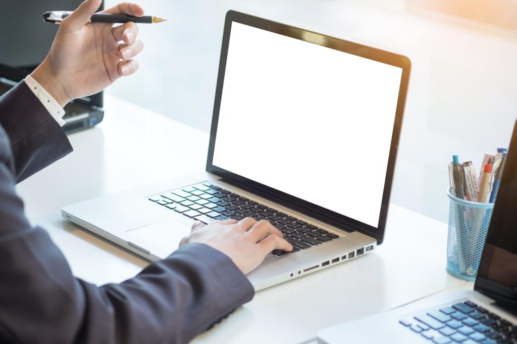 Person tippt an Laptop, andere Hand hält Stift, außerdem Stiftebehälter und weiterer Laptop am weißen Tisch,Qualität auf Schienen Bahnlogistik GmbH